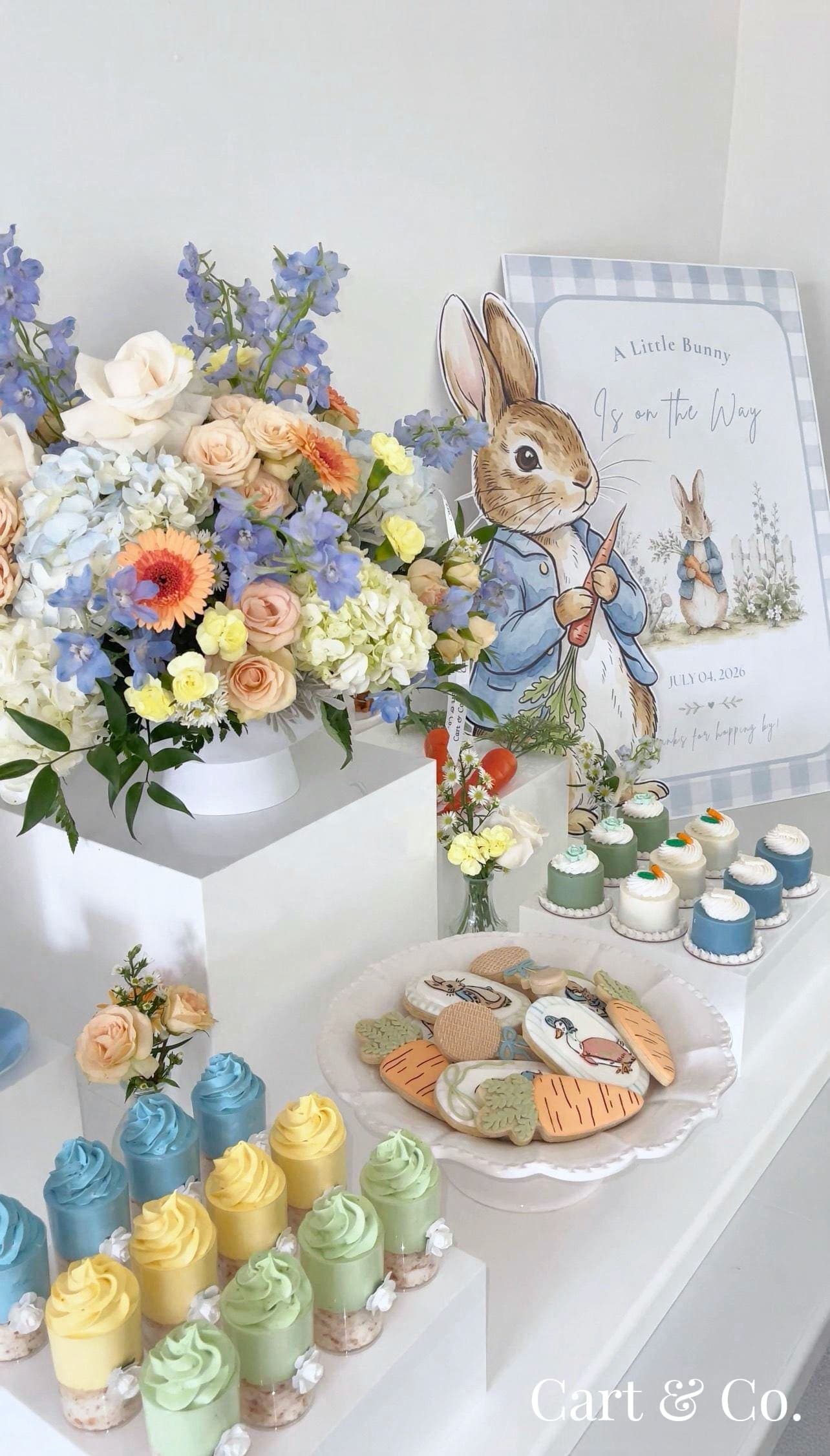 Peter Rabbit-themed dessert cart outdoors under a tree, styled with a pastel floral arrangement, a storybook and 'A Little Bunny Is on the Way' signage